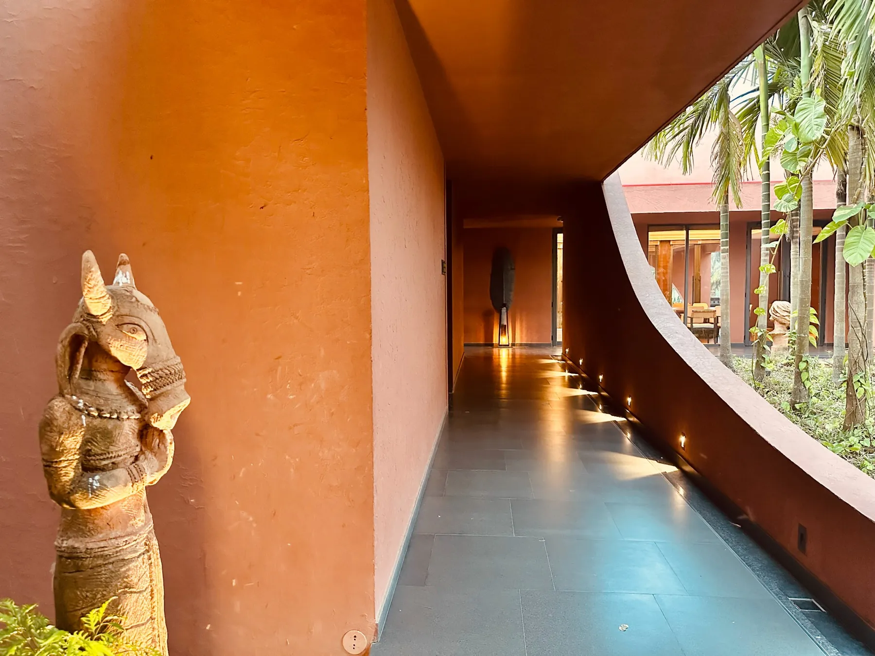 Terracotta passage with a sculpture and a curved opening towards the planted courtyard at Devasom, Kolkata